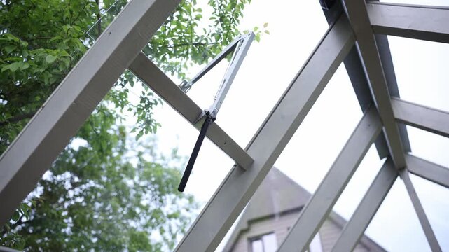 Upward view shows vent opener arm, hinge, and black lever on tempered glass roof. Subtle vent and leaf motion suggest airflow in a residential garden.