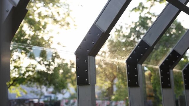Close up shows slanted wooden rafters, black metal brackets with rivets, and clear tempered glass. Sunlight at a low angle creates a starburst and backlit dust.