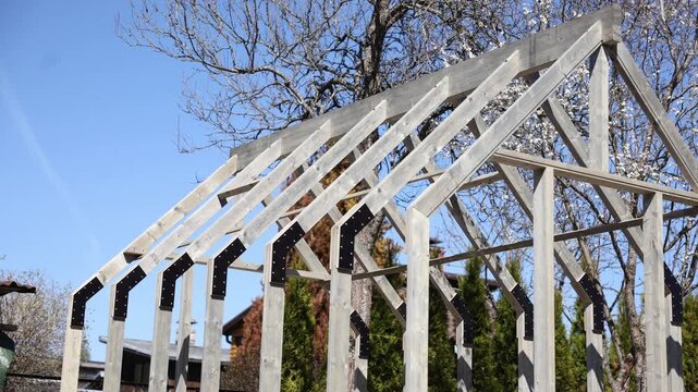 Unfinished wooden greenhouse frame shows triangular rafters, black bolted connector plates, and horizontal braces as the camera pans laterally in bright daylight.