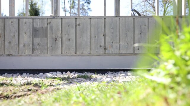 Low angle view shows weathered wooden panels, tempered glass panes, and a gravel drainage strip. Gentle lateral camera movement and plant motion occur in daylight.