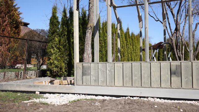 Timber greenhouse frame under construction with vertical wainscoting, white stone bed, metal ladder, rope secured tree trunk, and slow pan in natural daylight.