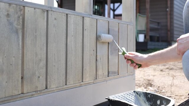 Close up shows a person priming vertical weathered wooden boards with a small roller. Even lateral strokes run above a concrete plinth and gravel strip in daylight.