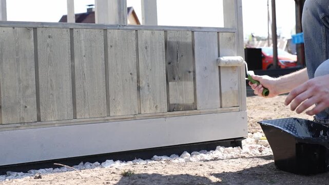Close view of weathered wood panels and tempered glass as one DIY worker rolls primer on lower trim. Gravel bed and paint tray sit at base in daytime.