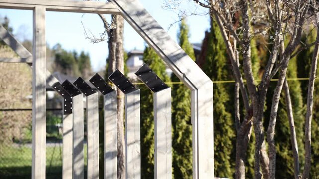 Construction of a wooden greenhouse frame in a backyard, with black steel roof brackets on posts, precise joinery, repeated bays, pans and focus pulls in daylight