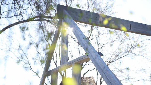 Close up view shows triangular timber frame, mortise and tenon joints, screw holes, and weathered grain in bright spring daylight with subtle leaf motion blur.