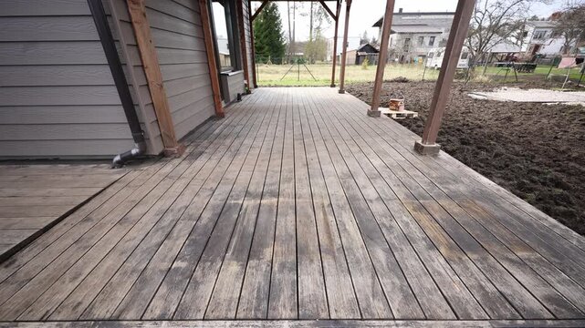Wide angle view shows long decking boards, vertical posts, metal downspout, and a pallet with paint cans on a porch. Overcast light highlights grooves and stains.