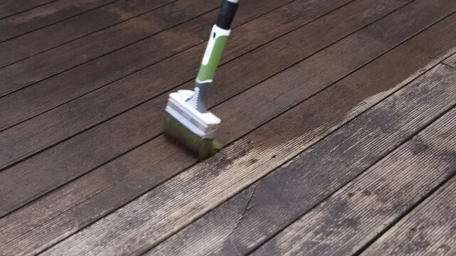 Green handled brush sweeps stain across weathered hardwood planks, showing darker treated grain, brisk motion, subtle moisture, and daylight outdoor setting.