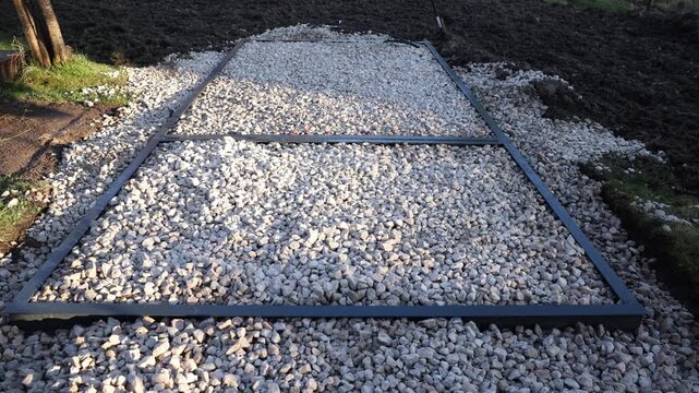 Rectangular steel frame sits in loose crushed stone as crossbars form a grid. Hand tools and a cable rest nearby. A tree trunk and turned soil border the base.