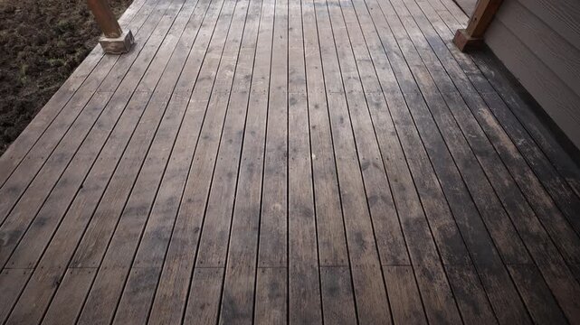 Parallel deck planks with dark circular stains and grooves run to the edge, flanked by square base posts and siding, dirt and turned earth suggest recent use in daylight