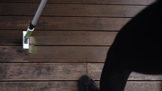 Top down view shows brisk scrubbing of weathered wood planks with green handled brush, black sneaker at edge, sweeping strokes upper right to lower left in daylight.