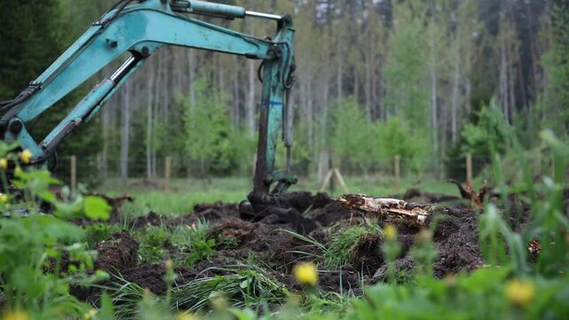 Turquoise excavator bucket digs turf, lifts sod and roots as soil falls. A rotted log lies nearby, wire fence runs by thin trunk trees in a rural field.