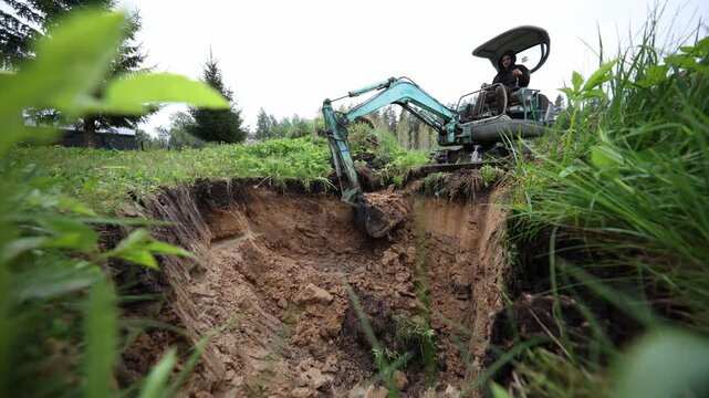 Turquoise mini excavator digs and lifts clay from a deep pit as earth falls. Operator in dark hoodie works near conifer trees and a small fence under overcast light.