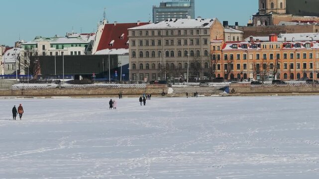 Riga Old Town embankment and Radisson Blu tower stand beyond the frozen Daugava as walkers cross packed ice, snow roofs and a large church tower rise in crisp daylight.