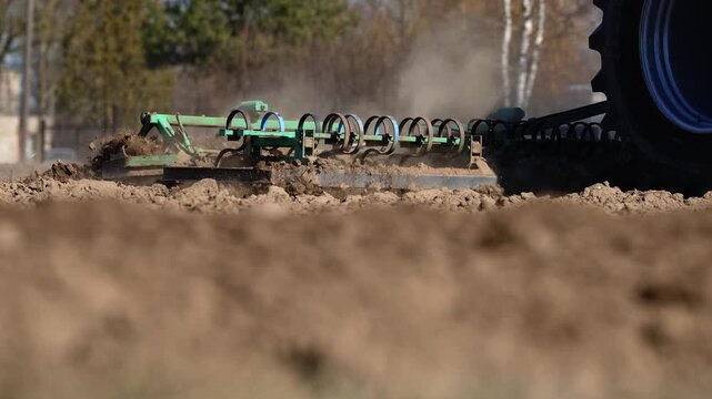 Low angle view shows a green coil spring harrow pulled by a tractor tire cutting dry soil, clods and dust fly rearward, tight focus, warm tones, high speed detail