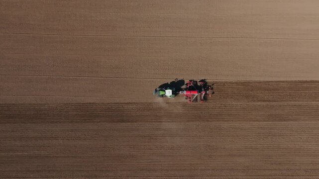 Aerial view shows a green tractor towing a red multi row planter across a brown field, moving in straight passes and carving even rows with a faint dust trail.
