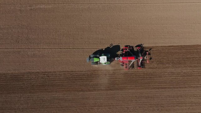 Aerial tractor with green cab pulls red seeder across brown field, forming parallel furrows as dust rises. Daylight, warm side light, graphic top down framing.