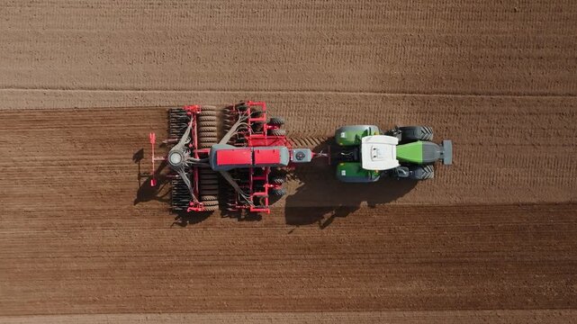Drone overhead view shows green tractor towing large red seeding implement across brown field, making parallel furrows, long shadows suggest seasonal planting