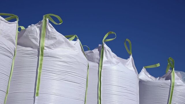 Large white woven sacks with neon lifting straps rise from a low viewpoint, crisp weave detail and vertical seams visible, slight strap motion in bright daylight.