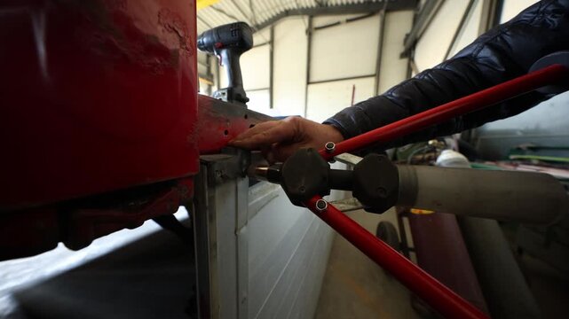 Close view shows clamp aligning a metal rod into a hinge as a drill rests on the rail. Camera tracks laterally in a large workshop with trusses and carts.