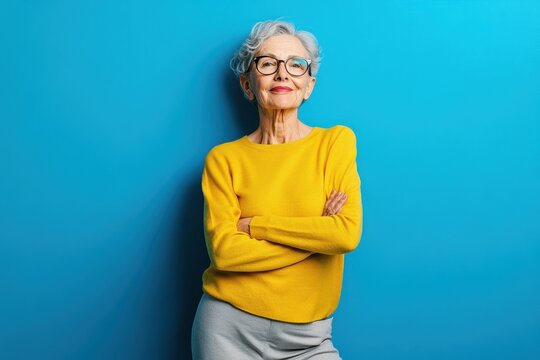 Confident senior woman with silver hair wearing glasses and a yellow sweater standing against a bright blue background