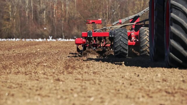 Low angle close up shows large tractor tires and red multi disc plow tilling brown field. Soil flips, dust sprays, and white gulls track progress near treeline.