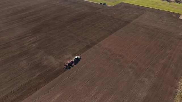 Aerial view of ploughed field with two tractors working in parallel. Long furrows, dust plumes, parked vehicles, and low sun cast elongated shadows show motion.