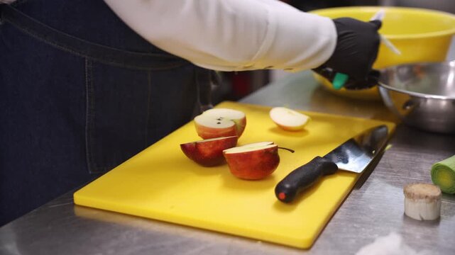 Hands in black nitrile gloves slice apple wedges on yellow cutting board next to chef's knife and steel bowl. Do not use a specific location and what is in title. Steady motion under bright light.
