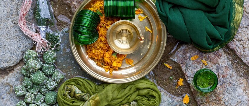 Polished brass thali with green glass bangles and marigold petals for Hariyali Teej, arranged on damp weathered stone with copy space for greetings