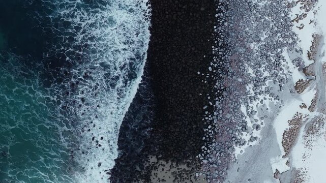 Top down aerial of dark Arctic beach bordered by surf and snow, drone establishing background, ascend reveal of rocks and beach