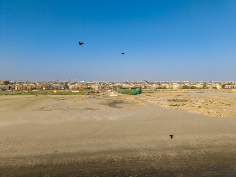 Aerial view of Sea-View Beach with sandy terrain, urban buildings in the distance, and birds flying under a clear blue sky in Karachi, Sindh, Pakistan.