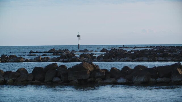 Calm tracking shot of a traditional fisherman standing on dark rocky breakwater piers at dusk, overlooking the quiet sea entrance of the Venetian Lagoon, Eraclea, Italy