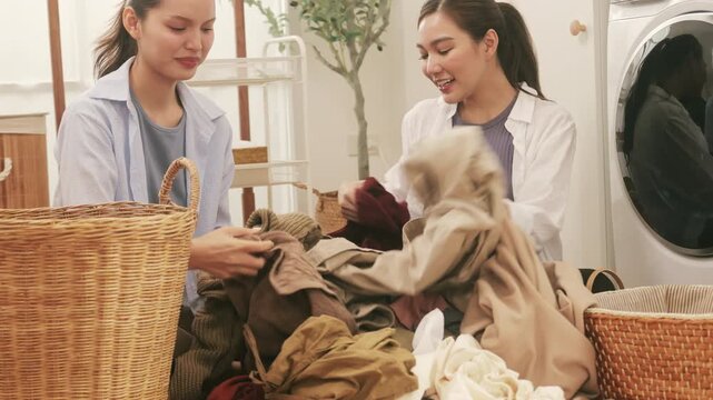 Two young Asian women cheerfully sorting clothes for laundry on the floor at home.
