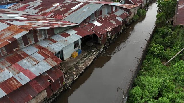 An aerial view showing a poor riverside shantytown with rows of rusty corrugated tin roofs lining a muddy, brown canal in Indonesia.