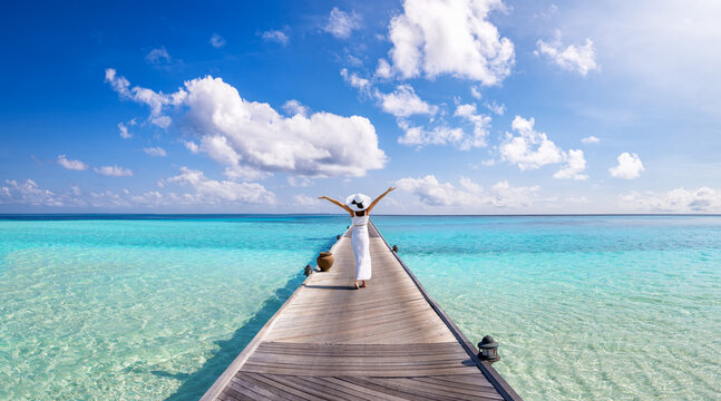 anoramic view of a happy woman in a white summer dress walking over a long pier over turquoise ocean in the Maldives islands as a travel concept