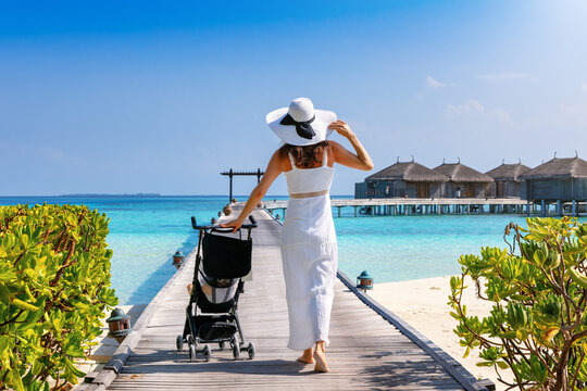 A elegant woman in a white dress walks over a pier in the Maldives islands pushing her baby in the stroller as a concept for traveling with a baby