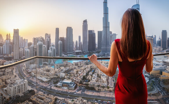 Rear view of a beautiful woman in a red dress looking from her balcony at the urban skyline of Downtown Dubai, UAE