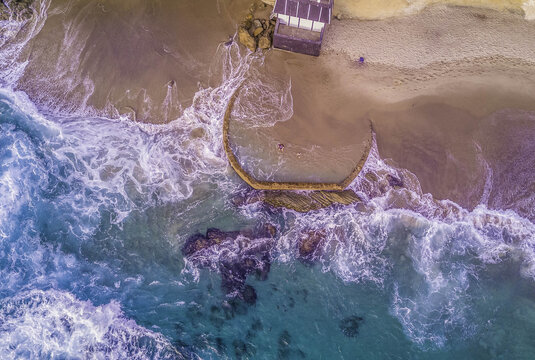 Aerial view of Victoria Beach featuring the iconic circular stone pool, turquoise waves crashing against rocky shorelines, and sandy beach in Laguna Beach, California, United States.