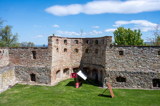 Visitors walk around the Boskovice Castle ruins in Czechia. Green grass covers the area, and trees are visible in the background. The sun shines bright in the sky.