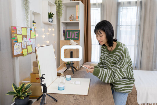 Young Online Seller carefully arranges skincare products and captures photos with her smartphone and ring lights setup in Home Studio Workspace
