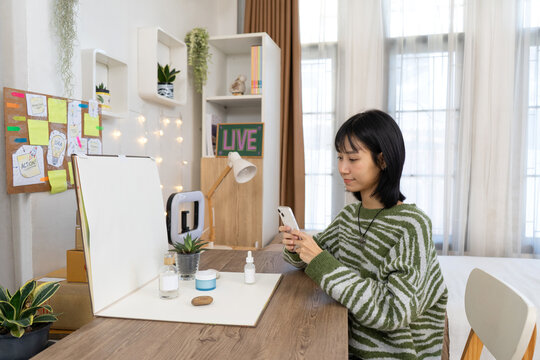 Young Online Seller carefully arranges skincare products and captures photos with her smartphone in Home Studio Workspace