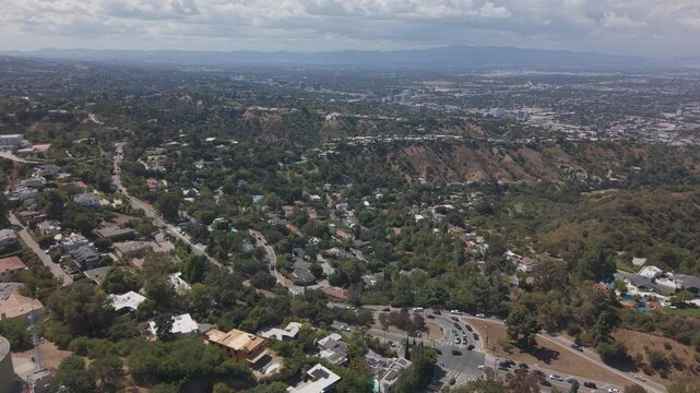 Hillside water tank and homes dot chaparral slopes above Sherman Oaks, with San Fernando Valley and distant mountains under cloudy skies in Sherman Oaks, California, USA. Aerial drone flyover shot