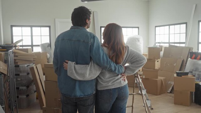 Woman and man hug with arms around waist amid stacked moving boxes and ladder in a building; hopeful new beginning.