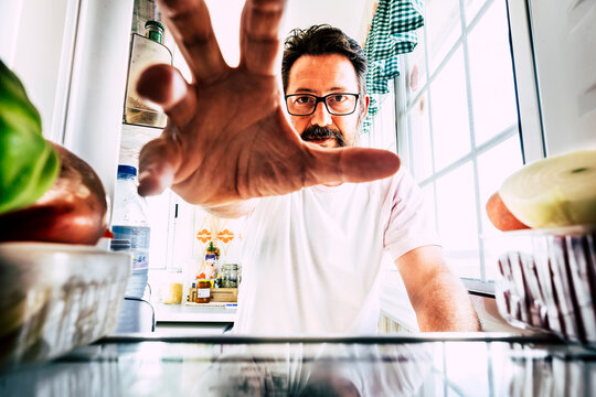 Man in glasses reaching into an open fridge in a bright kitchen, viewed from inside the refrigerator, conveying curiosity, hunger, morning routine and domestic life.