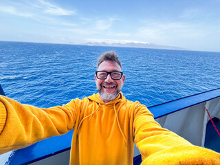Fototapeta premium Smiling man in yellow hoodie takes a selfie on a boat with deep blue ocean and distant islands, conveying travel, happiness and adventure on a sunny day at sea