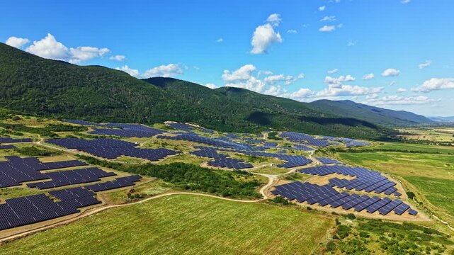 Rows of solar panels stretch across a green field near hills under a clear blue sky. This scene shows renewable energy efforts in a rural landscape.