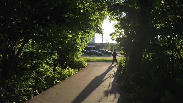 Tracking forward shot moving along a dark paved pedestrian path framed by lush green trees towards a sunlit city street