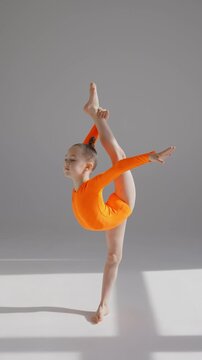 A little Caucasian girl gymnast in an orange leotard demonstrates her skills on a white background. 