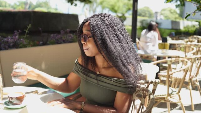 Young and beautiful black woman drinking water in a cafe and reading a book on a sunny and windy day