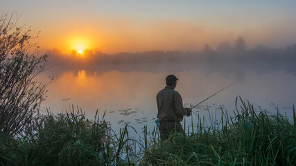 A fisherman stands amidst tall grass at dawn, casting his line into a misty lake as the golden sun rises © Александр Арендарь