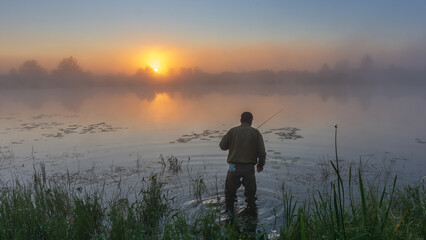 fisherman stands in the calm water at sunrise, surrounded by thick morning mist and the golden glowing sun © Александр Арендарь